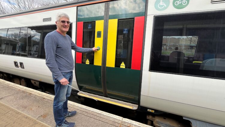 A male wearing dark glasses is standing on a train station platform, holding a white cane and pressing the door button on a train before he boards. The train door is marked with bright yellow and red panels for visibility, and symbols above the windows indicate spaces for wheelchair users and bicycles. Trees and a blue sky are visible in the background.