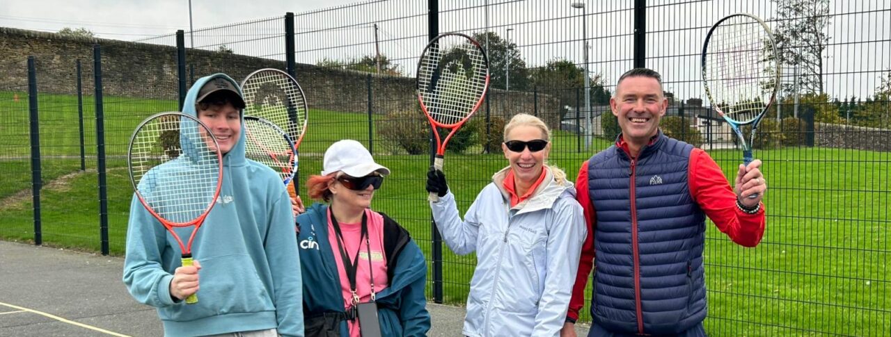 Three event participants and a sports coach stand on an outdoor court holding tennis rackets upright. They are dressed in casual sportswear and positioned close together. Behind them is a tall wire fence and a grassy area.