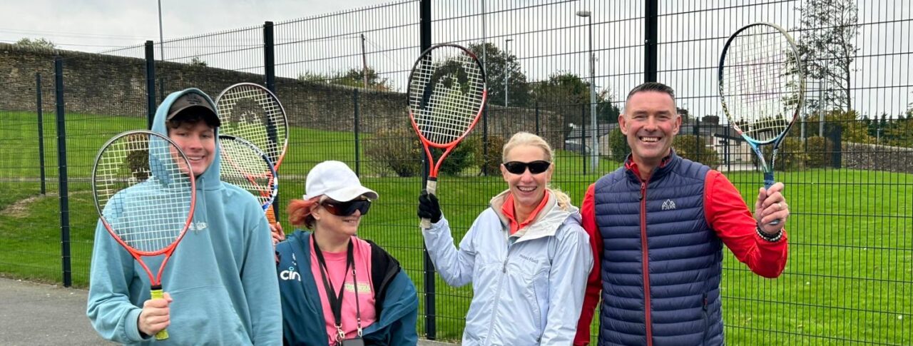 Three event participants and a sports coach stand on an outdoor court holding tennis rackets upright. They are dressed in casual sportswear and positioned close together. Behind them is a tall wire fence and a grassy area.
