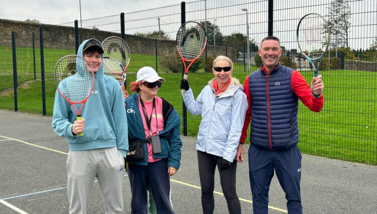 Three event participants and a sports coach stand on an outdoor court holding tennis rackets upright. They are dressed in casual sportswear and positioned close together. Behind them is a tall wire fence and a grassy area.