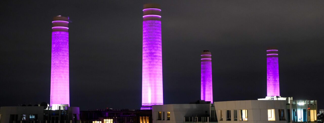 Arial photo of Battersea Power Station at night, with it's four chimneys lit up in purple.