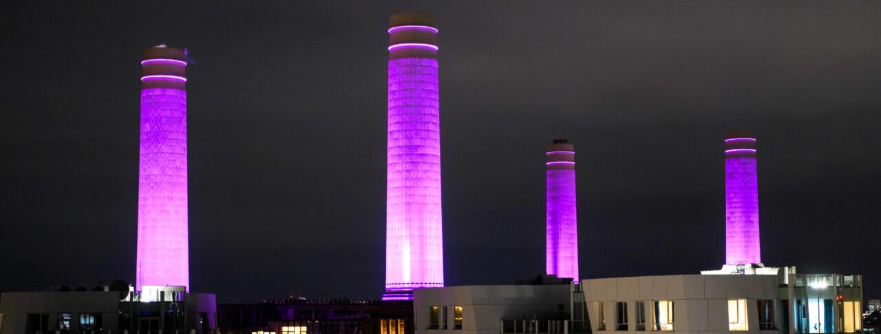 Arial photo of Battersea Power Station at night, with it's four chimneys lit up in purple.