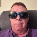 Headshot of Dale Durrant, Derbyshire SLC member. He is sitting on a dark grey sofa and wearing a purple shirt and sunglasses.