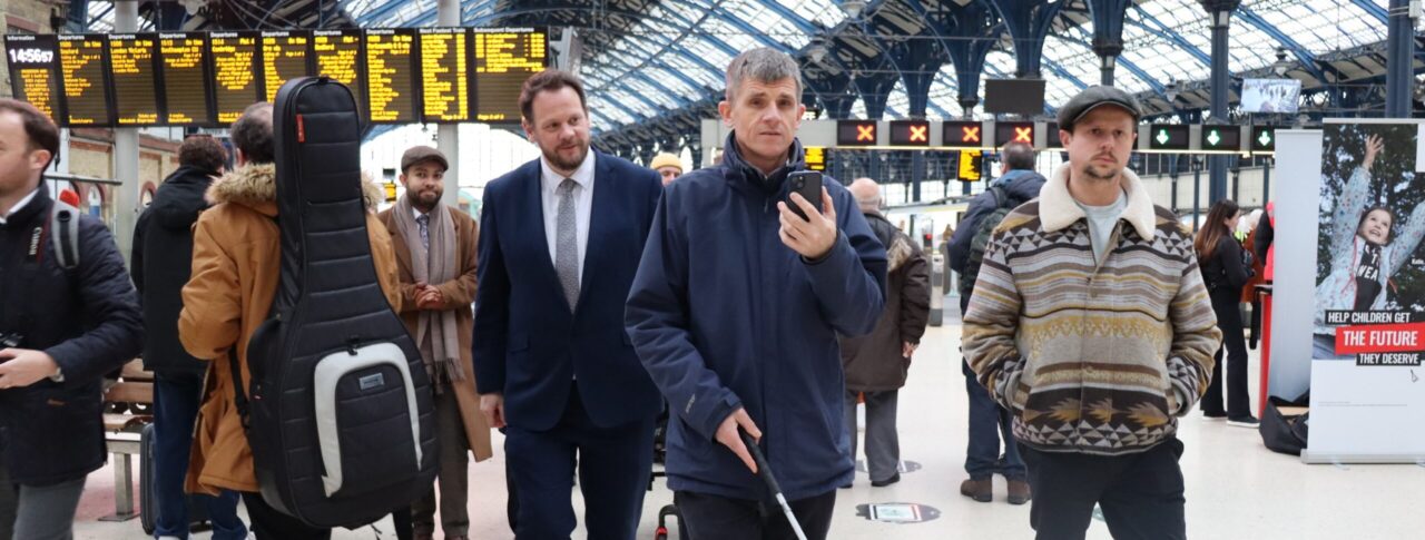 Dave Smith demonstrating the AIRA app inside a station to the Local Transport Minister in Brighton. He is walking holding his smartphone which he uses to speak to an AIRA operator. Transport Minister Simon Lightfoot walks behind him. In the background are crowds of people and passengers, and the stations electronic information boards.