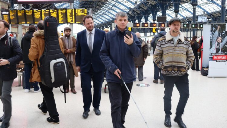 Dave Smith demonstrating the AIRA app inside a station to the Local Transport Minister in Brighton. He is walking holding his smartphone which he uses to speak to an AIRA operator. Transport Minister Simon Lightfoot walks behind him. In the background are crowds of people and passengers, and the stations electronic information boards.