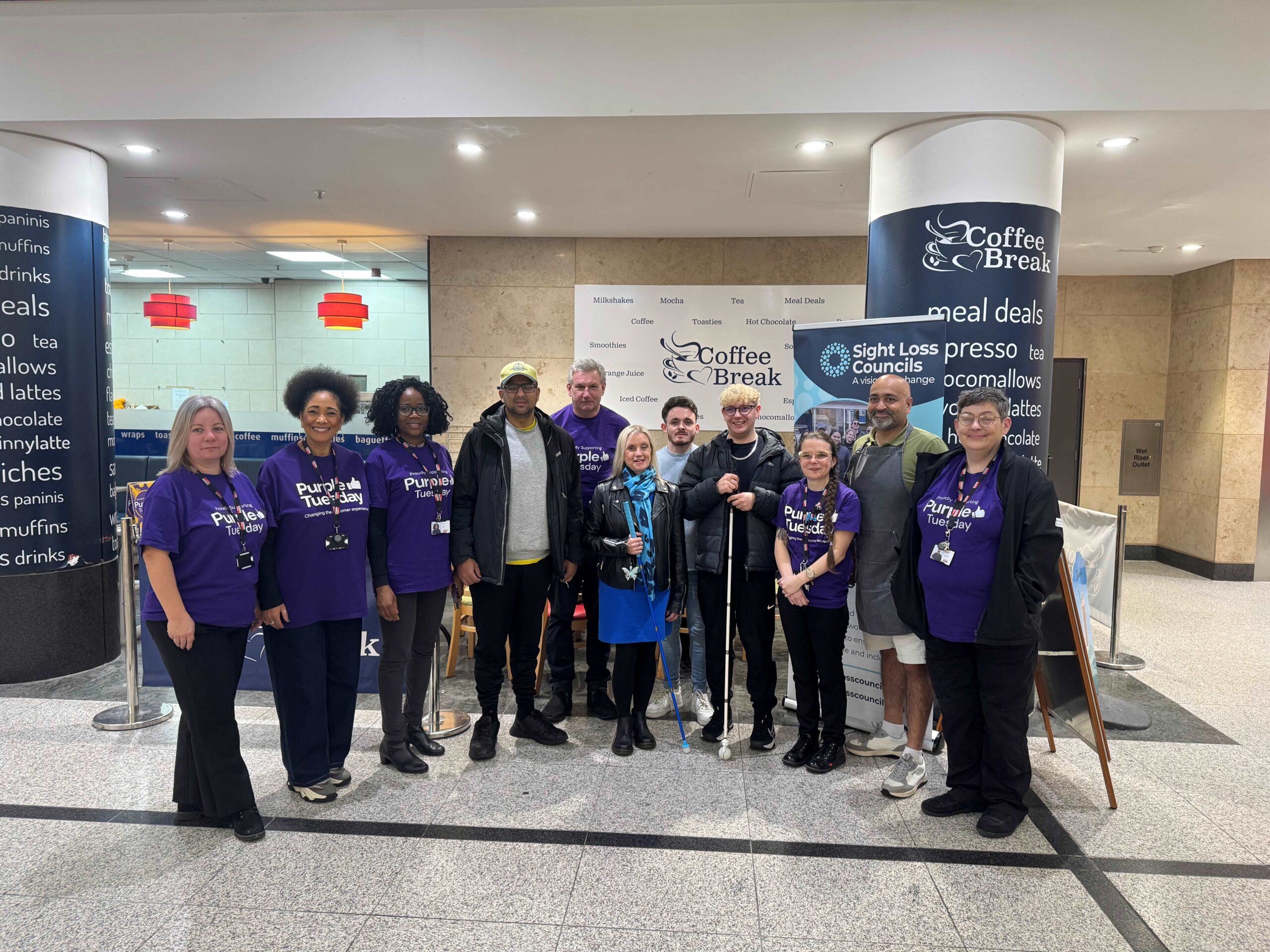 A group shot of Greater Manchester Sight Loss Council with staff from Manchester Arndale. Arndale staff are all wearing Purple Tuesday jumpers and hoodies. Everyone is smiling at the camera.