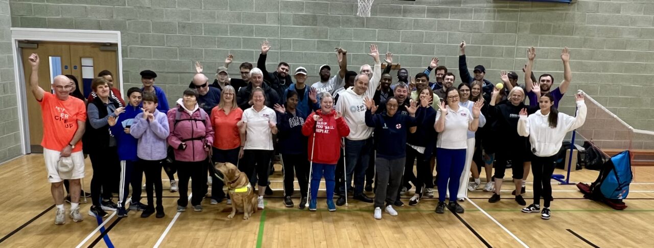 A group shot, taken in the sports hall, of participants, coaches, and staff after the SE London SLC Live Well, Get Active Day. Everyone is cheering at the camera, hands raised in the air.