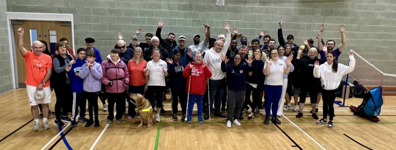 A group shot, taken in the sports hall, of participants, coaches, and staff after the SE London SLC Live Well, Get Active Day. Everyone is cheering at the camera, hands raised in the air.