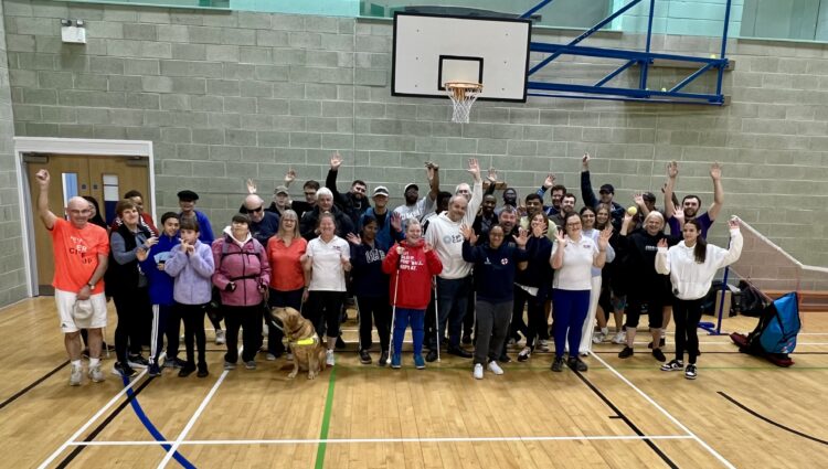 A group shot, taken in the sports hall, of participants, coaches, and staff after the SE London SLC Live Well, Get Active Day. Everyone is cheering at the camera, hands raised in the air.