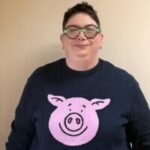 A headshot of Jemma Brown, Hampshire Sight Loss Council member. Gemma has short dark hair, and is wearing wide-rimmed, glasses. She is wearing a black jumper with a cartoon image of a pink pigs head on it, similar to Percy Pig!
