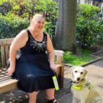 Leanne, London SLC member is sitting on a park bench with her guide dog. She is looking forwards, laughing.