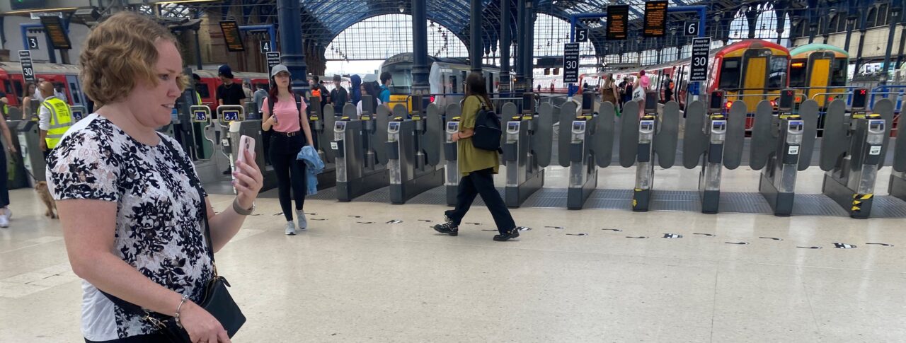 East Sussex SLC member, Linn, is being guided through Brighton train station whilst using the Aira app. The ticket barriers to the platforms are shown in the background.