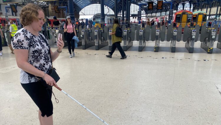 East Sussex SLC member, Linn, is being guided through Brighton train station whilst using the Aira app. The ticket barriers to the platforms are shown in the background.