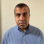 Headshot of Mohammed Mohsanali, who is standing against a cream wall. He is wearing a blue, gingham, shirt, and looking at the camera.