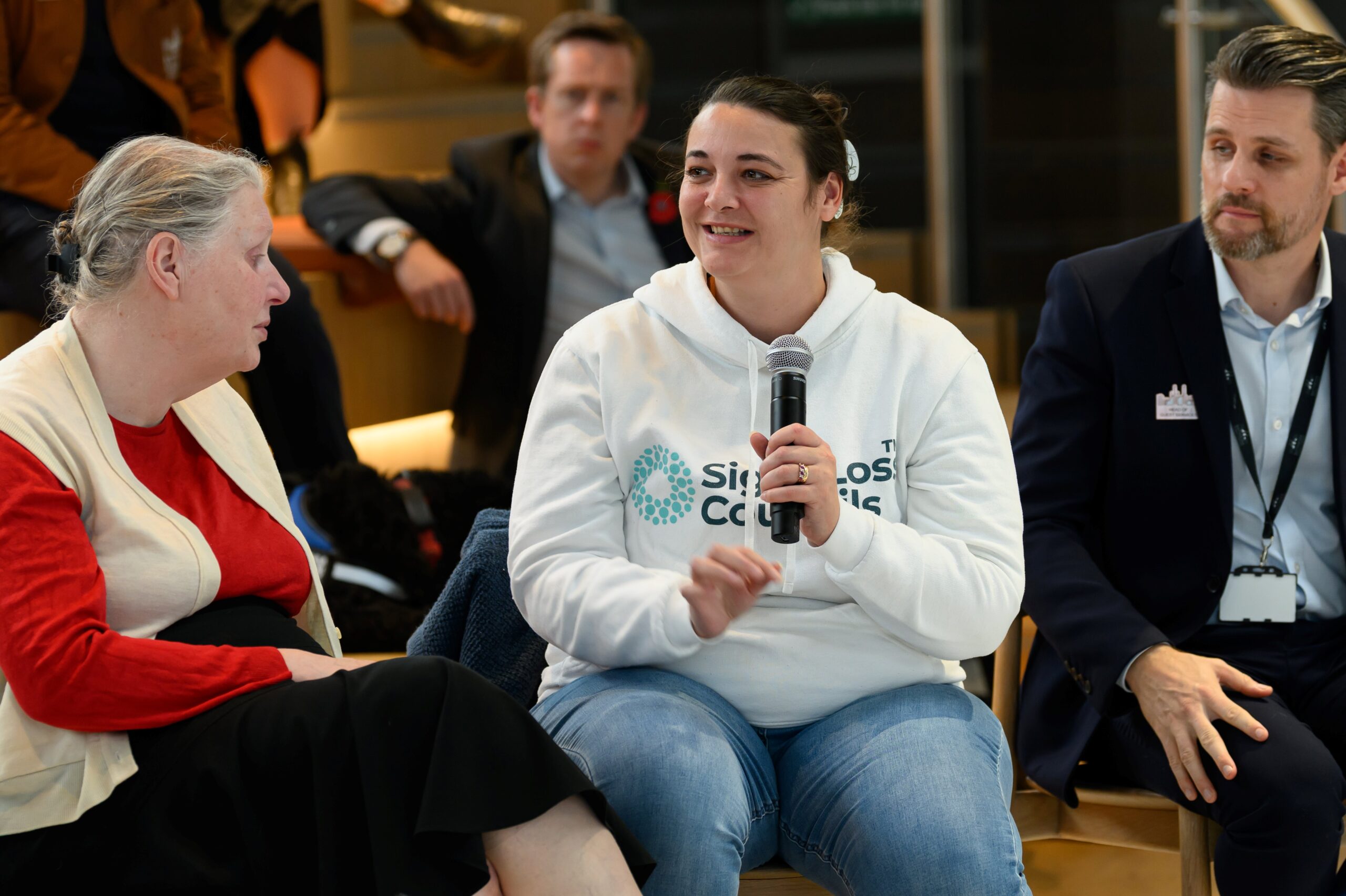 Nikki, South West London SLC member, is smiling as she talks into a microphone. She is sat next to fellow SLC member, Mary, and Head of Guest Services, JP.