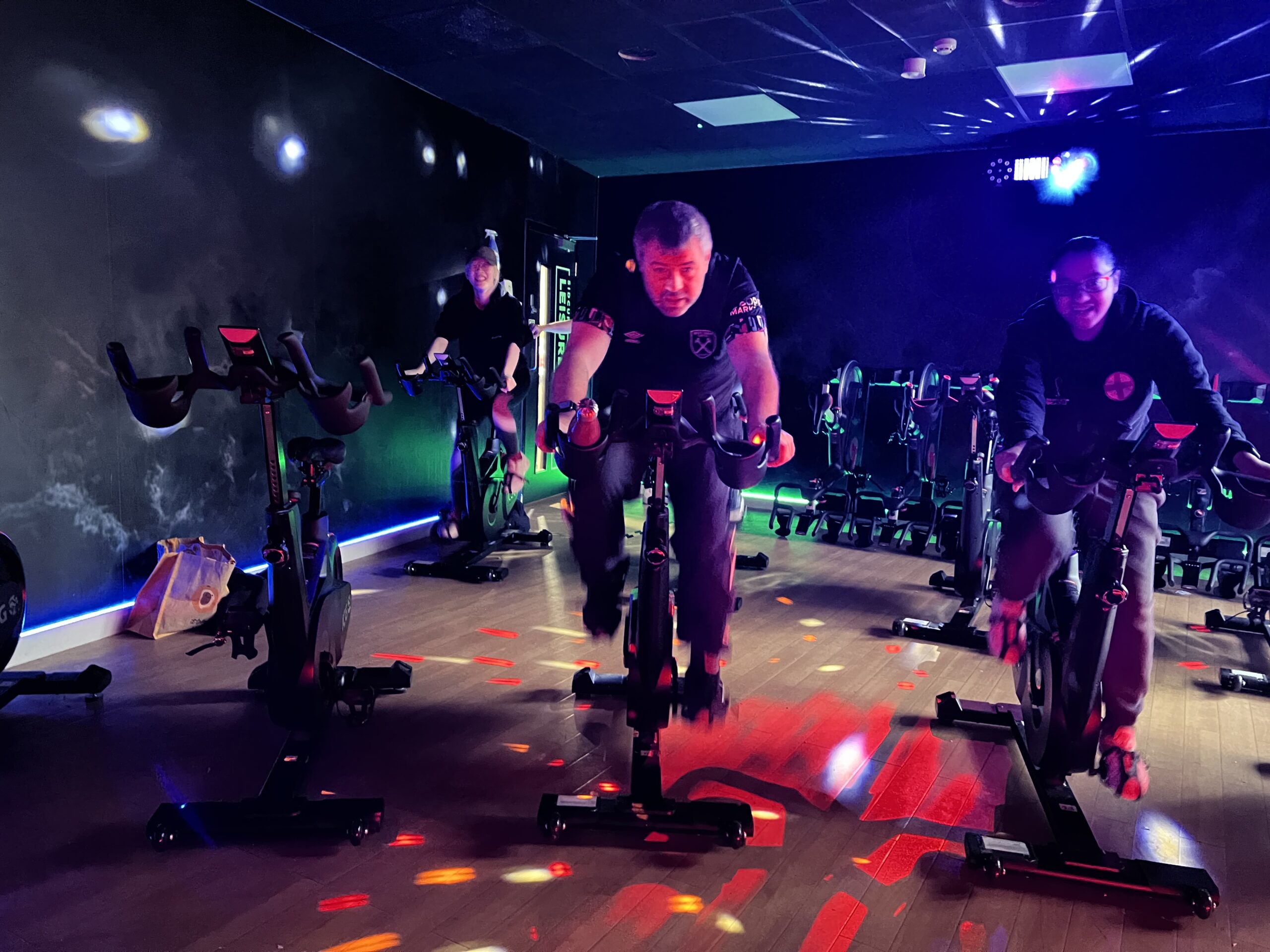 Steve Reed, SE London SLC member, participating in the spin session alongside fellow attendees. The studio is lit with multicolour lights, and he is glancing up at the camera.