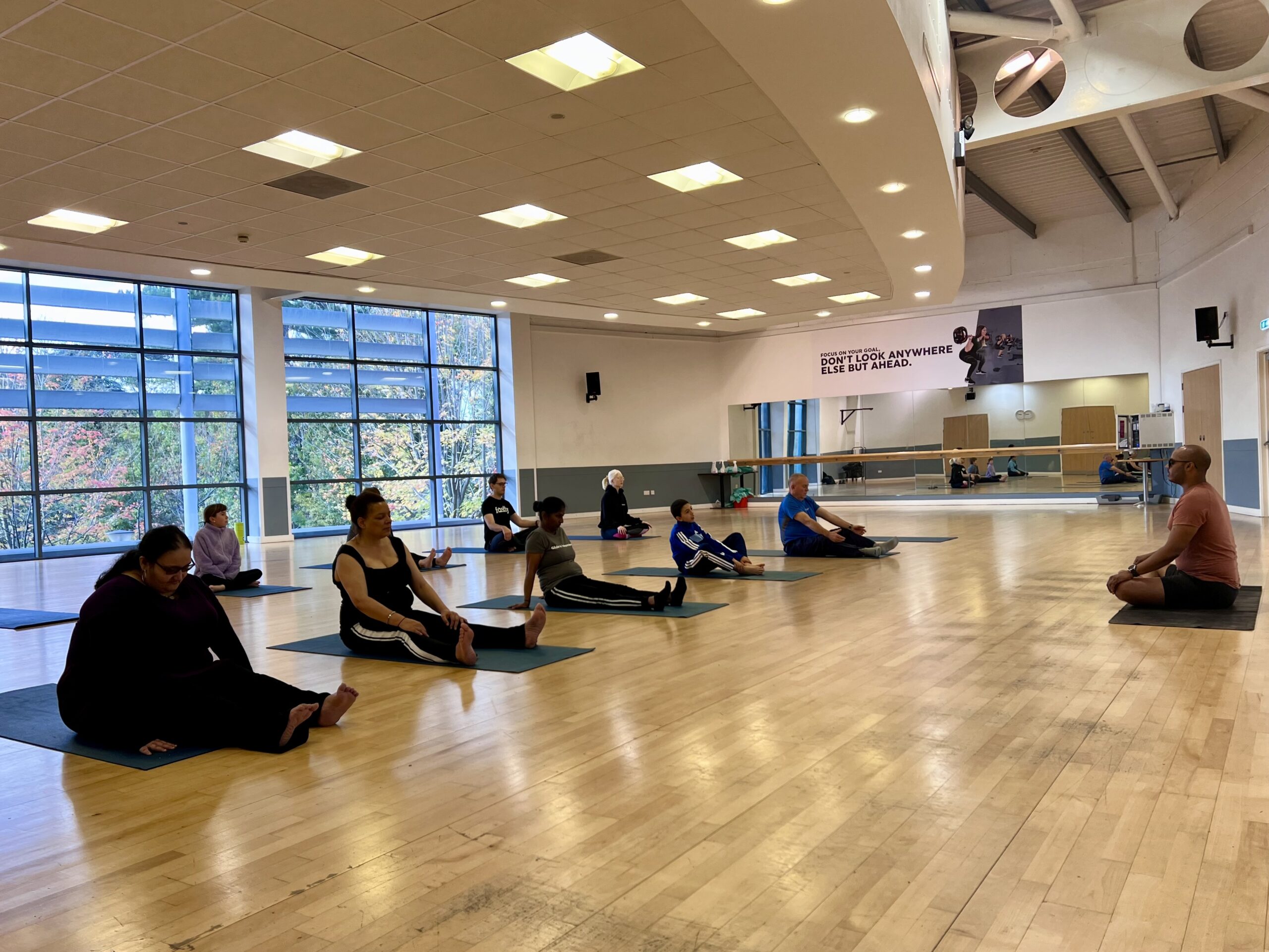 View across the dance studio where participants sit on yoga mats with their legs stretched in front of them. Ashfaq, the VI Yoga Instructor, sits at the front of the class on his mat with legs crossed, sitting tall.