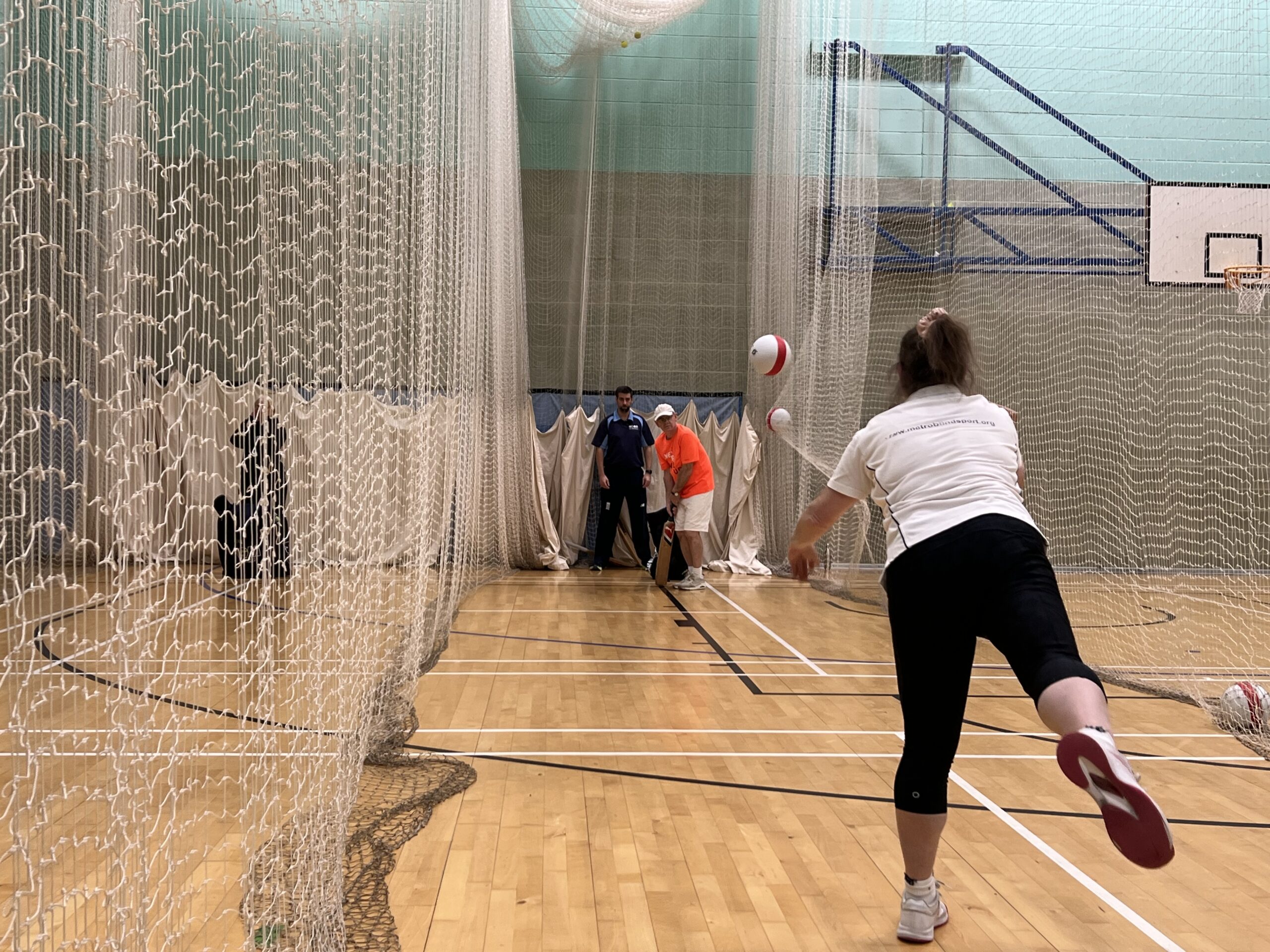 A cricket coach bowls to a visually impaired participant during a VI Cricket session. The photo is taken from behind the coach, mid-bowl, with the ball in mid-air between the bowler and batter. Floor-to-ceiling cricket nets hang on either side of them