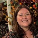 Amy Askew, Engagement Manager for East of England. Amy has long, dark hair and is weating a black dress with green abd pink flowers on it. Amy is stood in front of a large flower arrangement, made up of burnt orange and foliage.