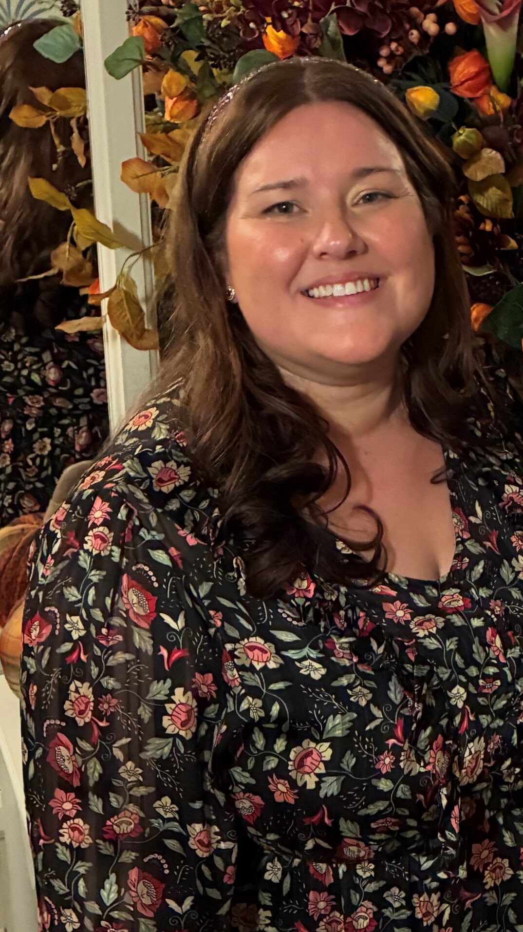 Amy Askew, Engagement Manager for East of England. Amy has long, dark hair and is weating a black dress with green abd pink flowers on it. Amy is stood in front of a large flower arrangement, made up of burnt orange and foliage.