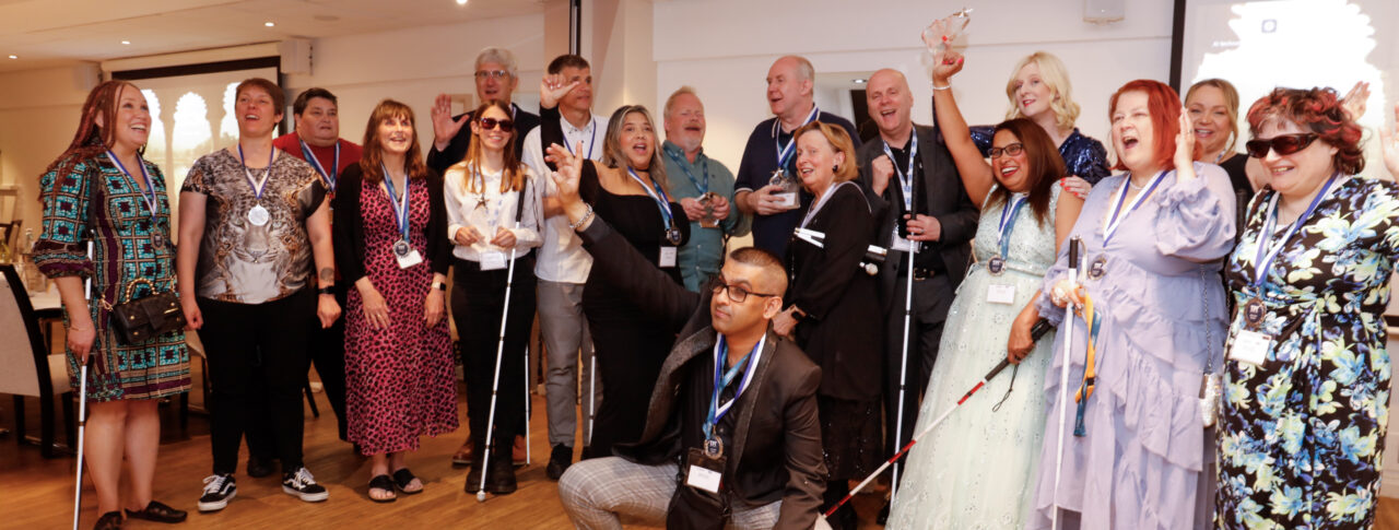 A group photo of eighteen people celebrating. They are smiling and cheering and many are wearing medals, with a few people also holding their canes. They are all smartly dressed and look happy. One man is crouched at the front with his arm in the air, celebrating.