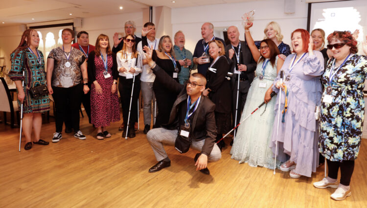 A group photo of eighteen people celebrating. They are smiling and cheering and many are wearing medals, with a few people also holding their canes. They are all smartly dressed and look happy. One man is crouched at the front with his arm in the air, celebrating.