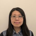 Head shot of Ching, SE London SLC volunteer. Cheer is standing against a beige wall, wearing clear framed glasses, smiling at the camera.