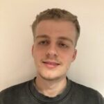 Headshot of Gledi Ndreu, a North London Sight Loss Council member in his twenties. He has short brown hair and is smiling at the camera. Gledi is wearing a black jumper and standing against a cream-coloured wall.