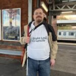 Lee Mercer, Essex SLC member, is stood on a train platform, holding his long cane. He is wearing his white Sight Loss Council hoodie, wearing a beige coat, and smiling at the camera. A train is in the background at another platform.