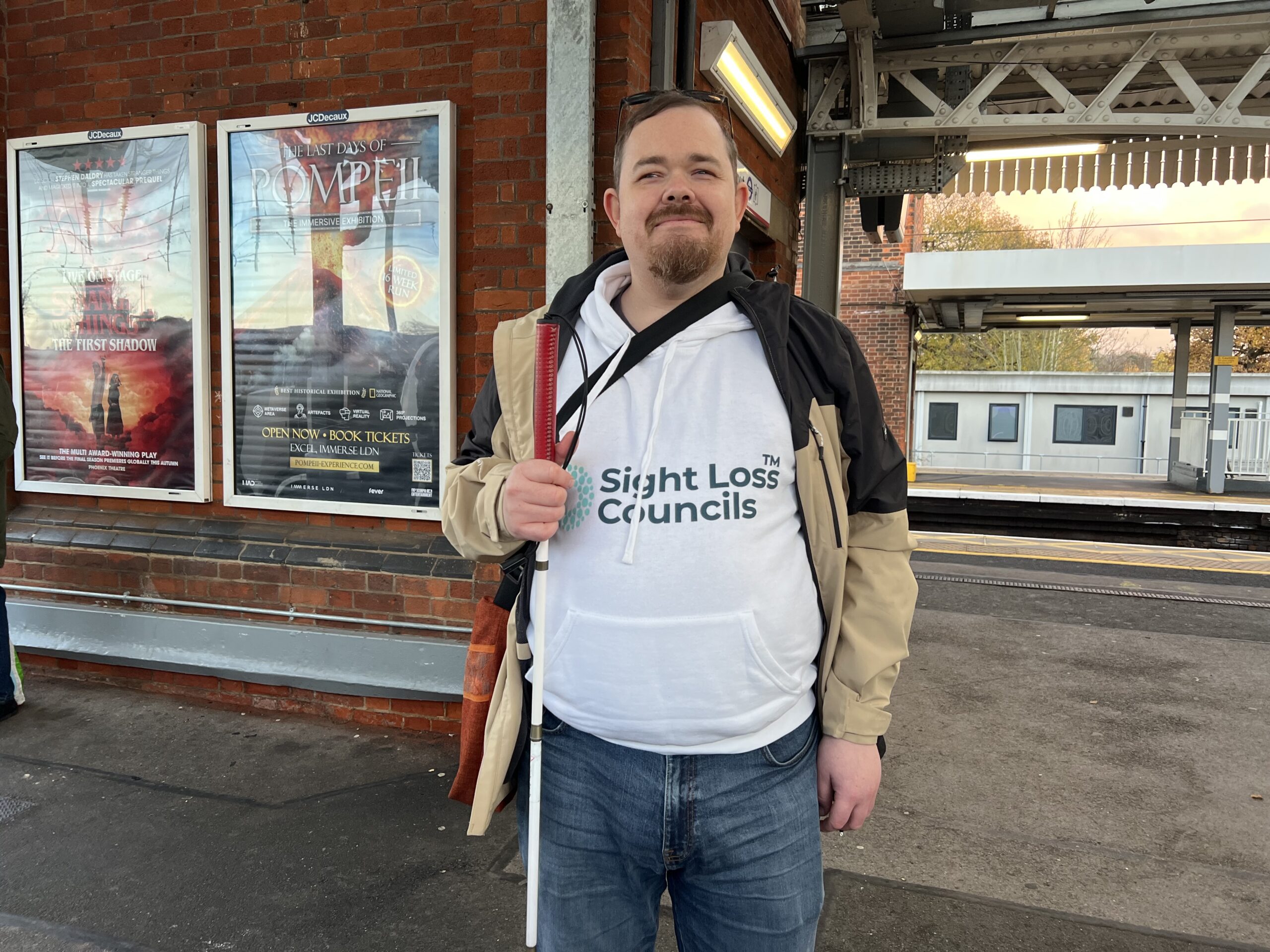 Lee Mercer, Essex SLC member, is stood on a train platform, holding his long cane. He is wearing his white Sight Loss Council hoodie, wearing a beige coat, and smiling at the camera. A train is in the background at another platform.