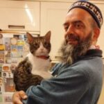 Kahl stands in a kitchen with cream cabinets, turned sideways to the camera. He is wearing a hat, has a grey beard, and is smiling while holding a grey and white cat with a red collar in his arms.