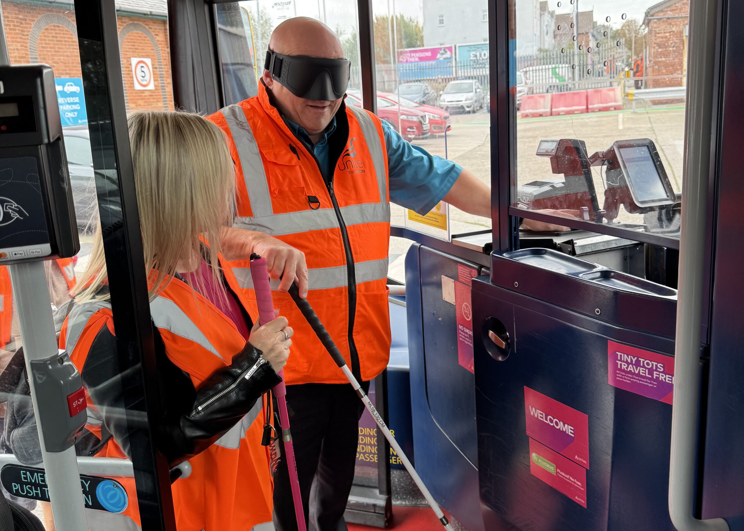 Kelly, Engagement Manager for North West England, is standing next to the bus cab with a male driver. He is wearing simulation glasses, holding a long cane, and trying to find the card reader as part of the bus driver awareness training session. Both are in orange hi-visibility jackets.