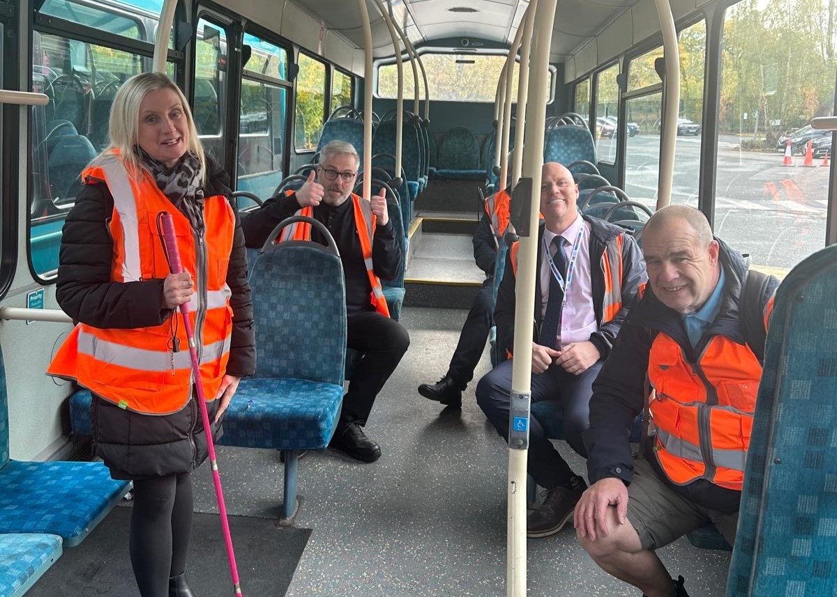 Kelly Barton, Engagement Manager for North West England, is standing on a stationary bus with her pink, long cane. Sat behind her is an Arriva bus driver with his thumbs up. On the opposite side of the bus, Neil Atherton, Bus Driver Instructor for the Northwest sits and smiles at the camera, with SLC meber, Mick, sitting in front of him.