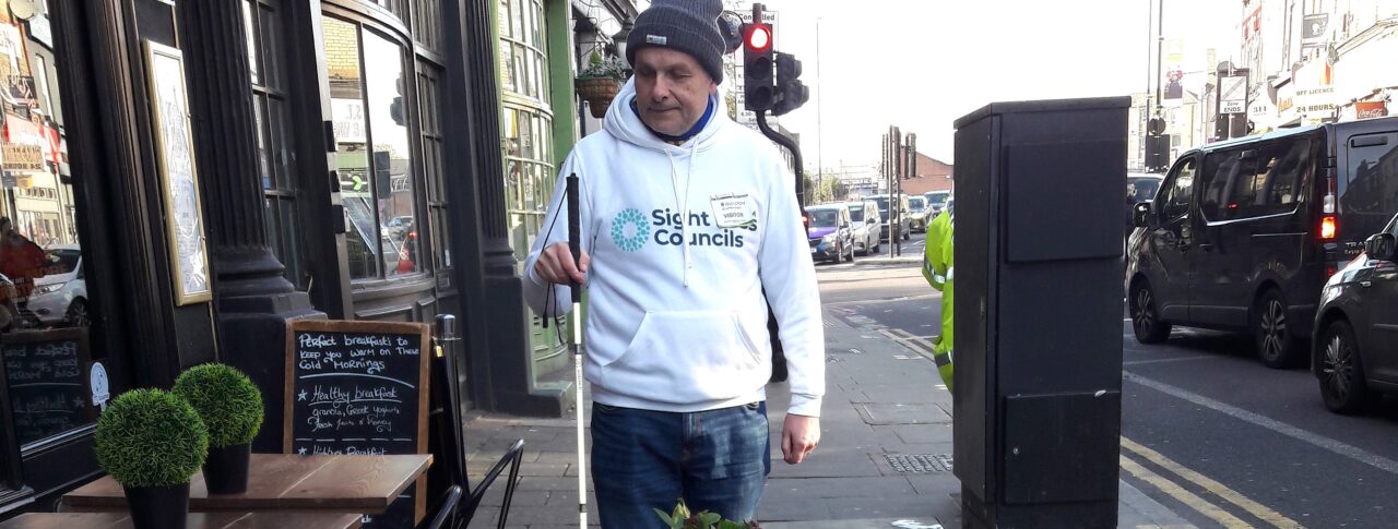 Liam O'Carroll, interim engagement Manager for London Sight Loss Councils, is walking along a pavement next to restaurant tables and chairs. He is holding his long cane, and about to walk into a planter, directly in front of him.