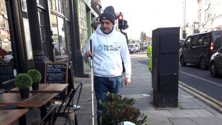 Liam O'Carroll, interim engagement Manager for London Sight Loss Councils, is walking along a pavement next to restaurant tables and chairs. He is holding his long cane, and about to walk into a planter, directly in front of him.