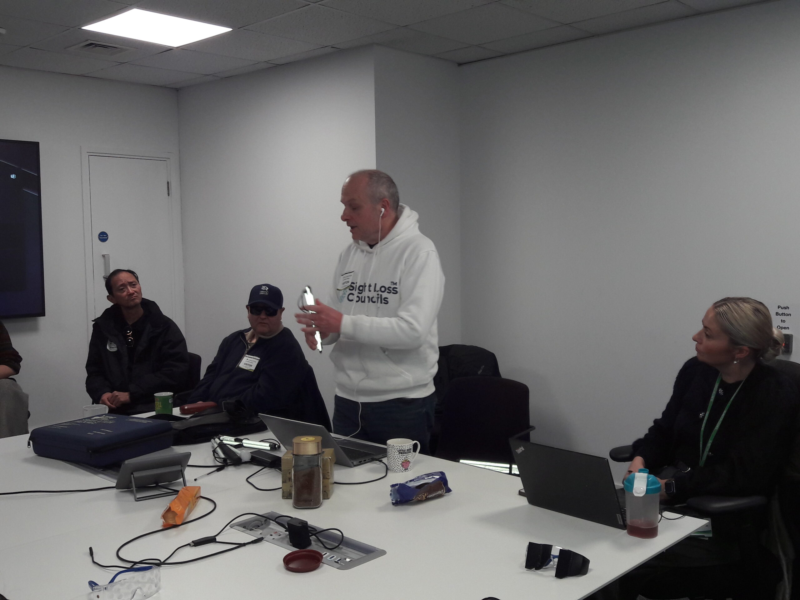 Liam O'Carroll, Interim Engagement Manager for London Sight Loss Councils is stood at a meeting table, holding a folded, cane. Attendees of the sim-spec walk are looking on as he demonstrates it's use.