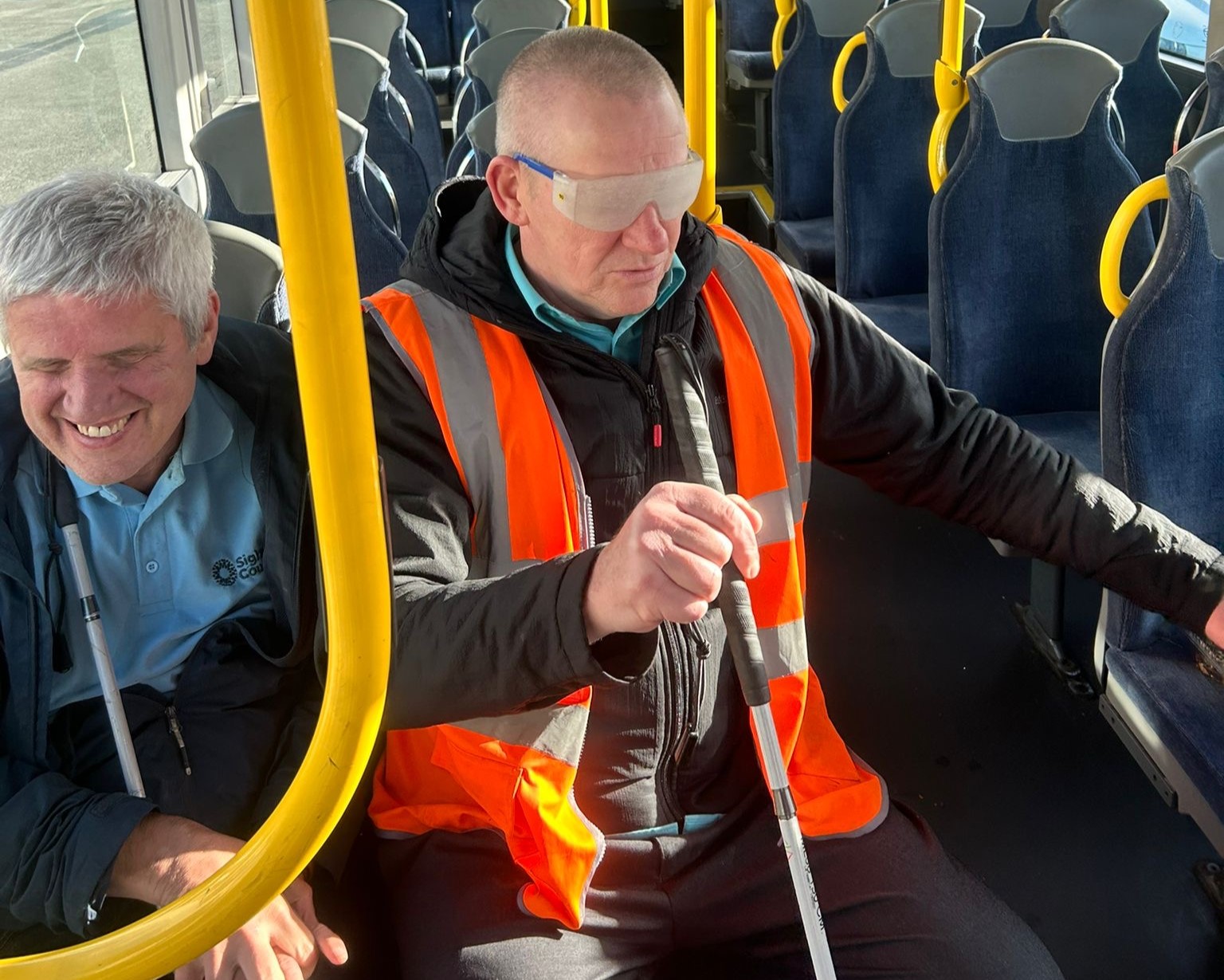 Mike Wright, Merseyside SLC member is sat on a bus alongside a bus driver during a Bus Driver Awareness session. Both men are in orange high-visibility vests and the driver, is wearing simulation glasses and holding a cane. Yellow handrails and rows of empty seats are visible in the background.