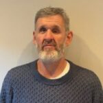 Photo of Richard Holmes, SW London Sight Loss Council volunteer. He has grey hair and a beard and is stood against a cream wall. He is looking forwards,smiling at the camera.