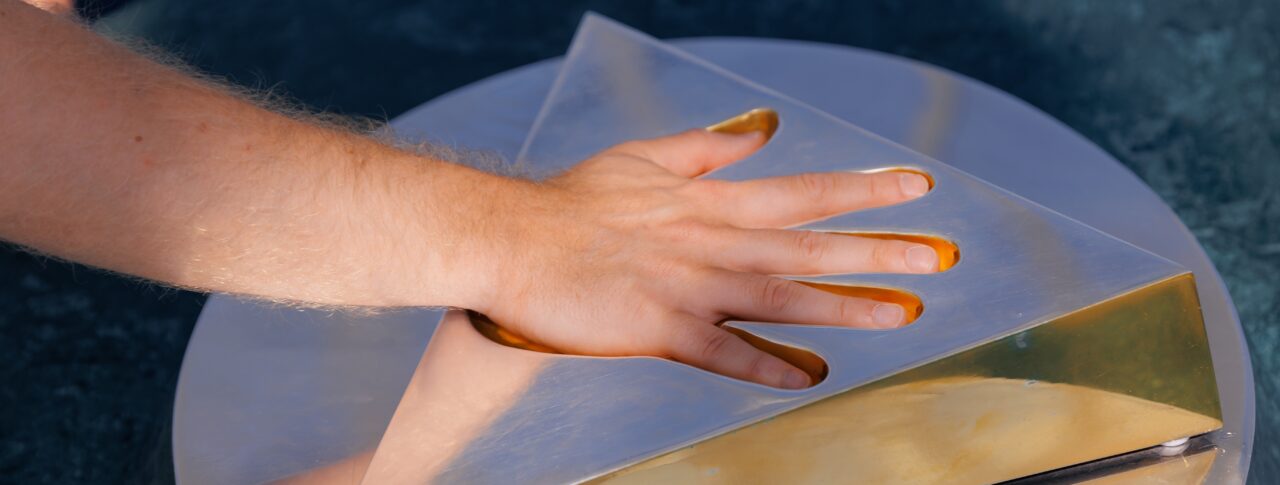 A hand pressing down on a metallic triangular sculpture with a reflective gold surface, placed on a circular base. The sculpture has a cut-out section that fits the hand, and the fingers appear to have a golden tint from contact with the surface.