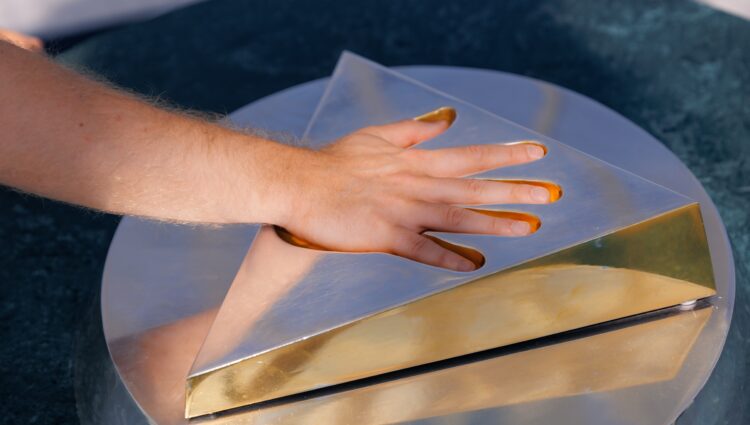 A hand pressing down on a metallic triangular sculpture with a reflective gold surface, placed on a circular base. The sculpture has a cut-out section that fits the hand, and the fingers appear to have a golden tint from contact with the surface.