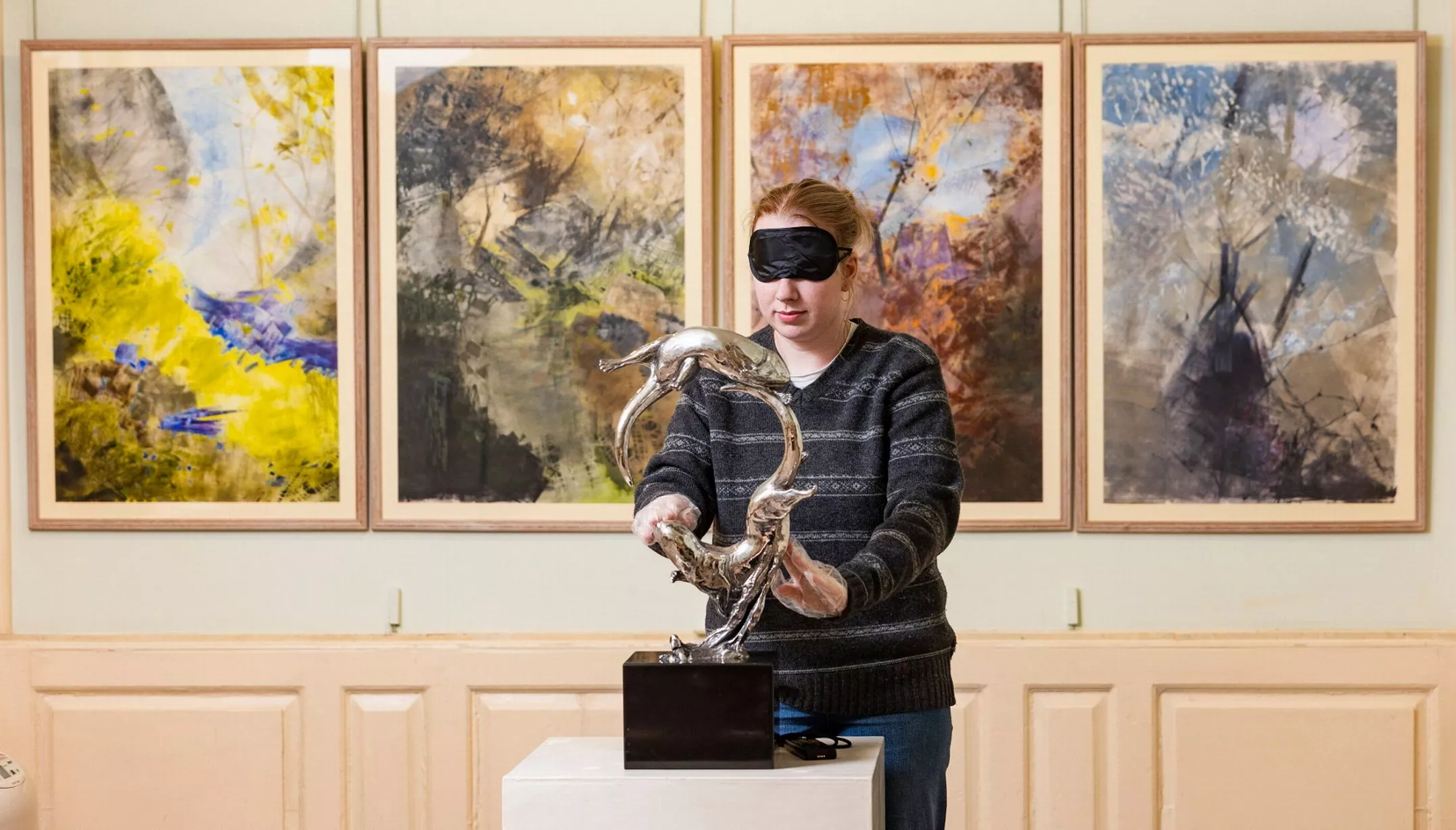 A blindfolded female stands in front of four, large, abstract paintings in earthy tones, mounted on a cream panelled wall. The person is feeling a metal sculpture on a white pedestal, which has two animals leaping in a circular formation.
