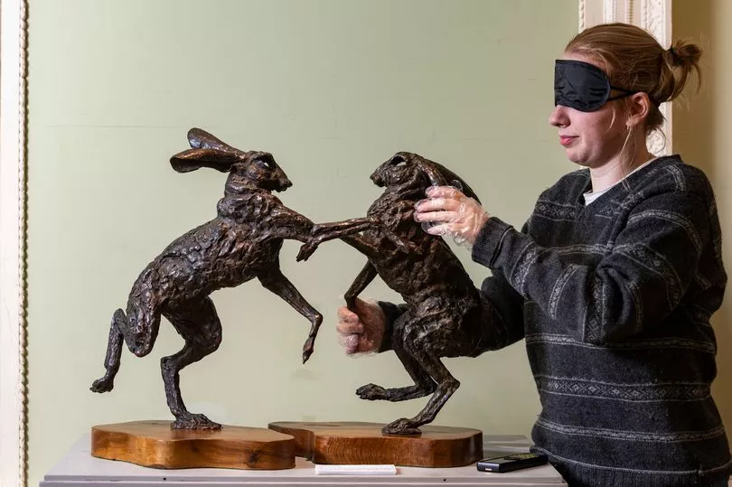 A bronze sculpture of two hares standing upright on their hind legs, in a boxing pose, mounted on separate wooden bases. The hares have elongated ears and detailed, textured, fur. One hare is leaning forward slightly and the other is pushing back. A female, wearing a blindfold, is exploring the sculpture through touch.
