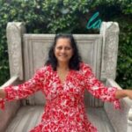 Vee is sat outside in a large wooden chair, against a large green hedge. She has shoulder length, dark, curly hair and is wearing a red and white patterned dress. She is smiling at the camera.