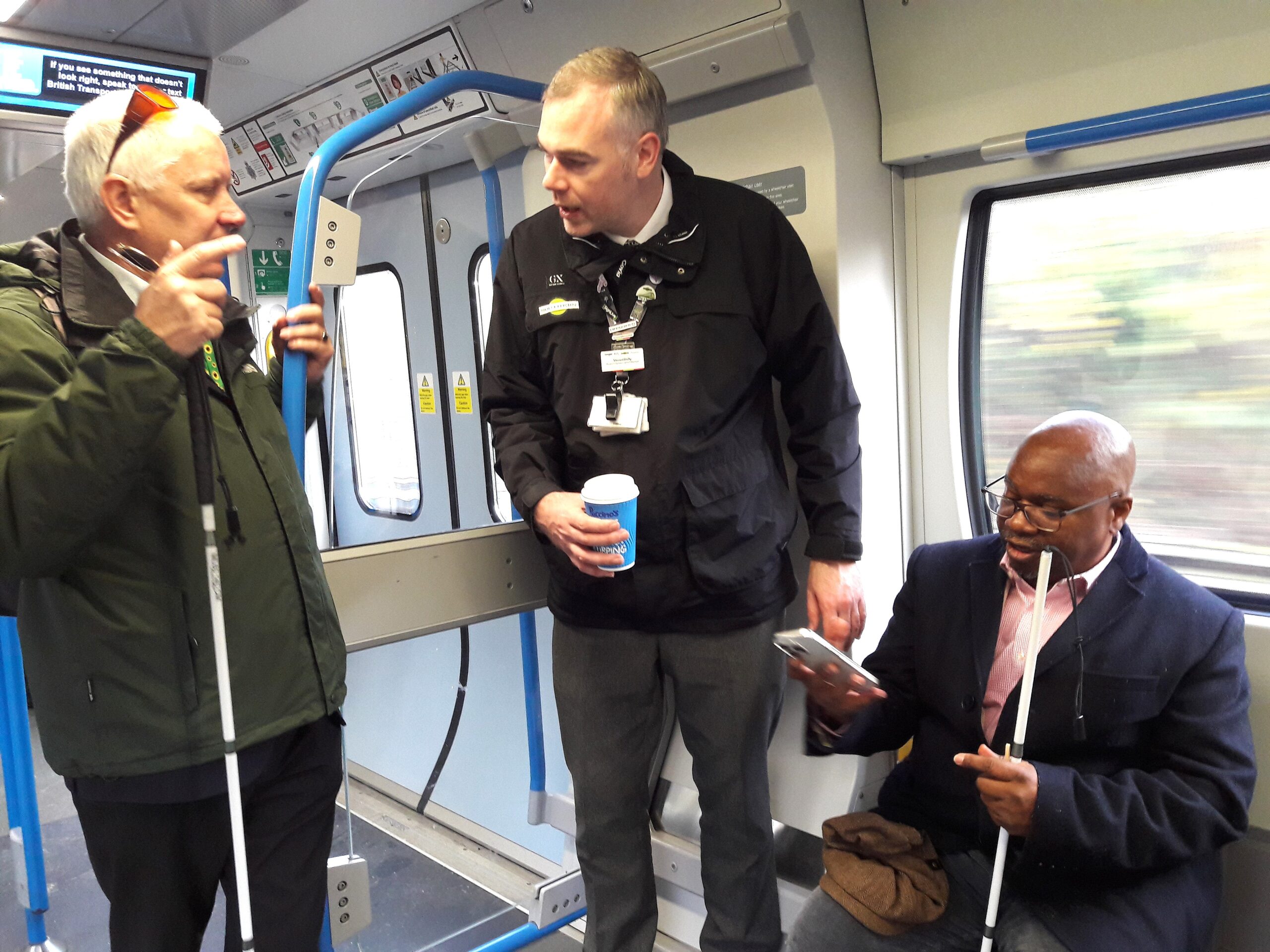 Vince, GTR staff member, speaks to an attendee on-board the train, with another sat down next to him. Both attendees are holding their long canes.