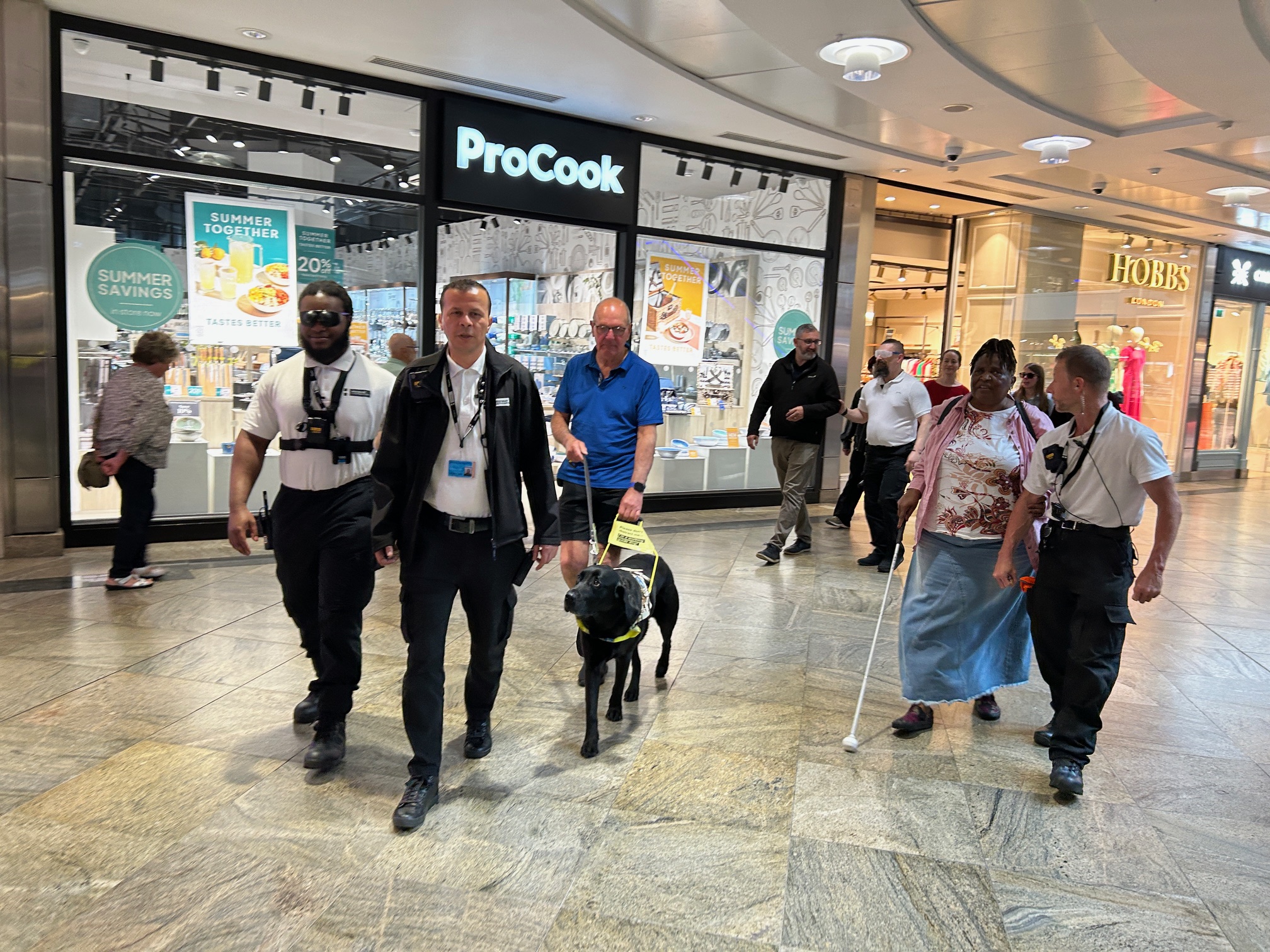 Members of Hampshire SLC with centre staff in West Quay Shopping Centre during the sighted guiding part of the VI Awareness session. The group are working in pairs, with one person wearing simulation glasses whilst guiding their partner.