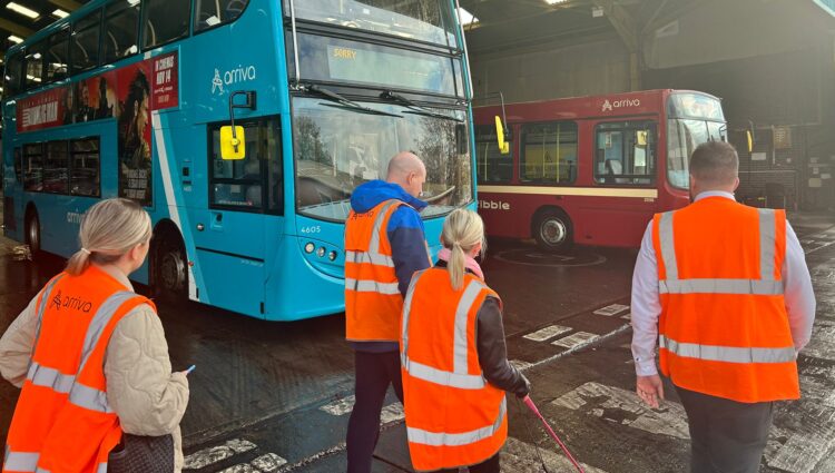Four people, wearing high-visibility vests, are walking in a bus depot towards two parked buses. One bus is turquoise with the Arriva logo, the other is red. Kelly, pictured in the centre, is walking with her long cane. The depot has a high rood and bright, overhead lights.
