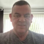 Selfie of Eric, Greater Manchester Sight Loss Council member. Eric ha short dark hair and is wearing a grey t-shirt. He is stood in front of a window which has white blinds drawn.