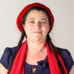 Photo of Caroline Devonport, West Yorkshire SLC member. She has long dark hair, tied in two plaits. She is wearing a dark top, a red scarf and red beret. She is stood against a white wall, smiling at the camera.