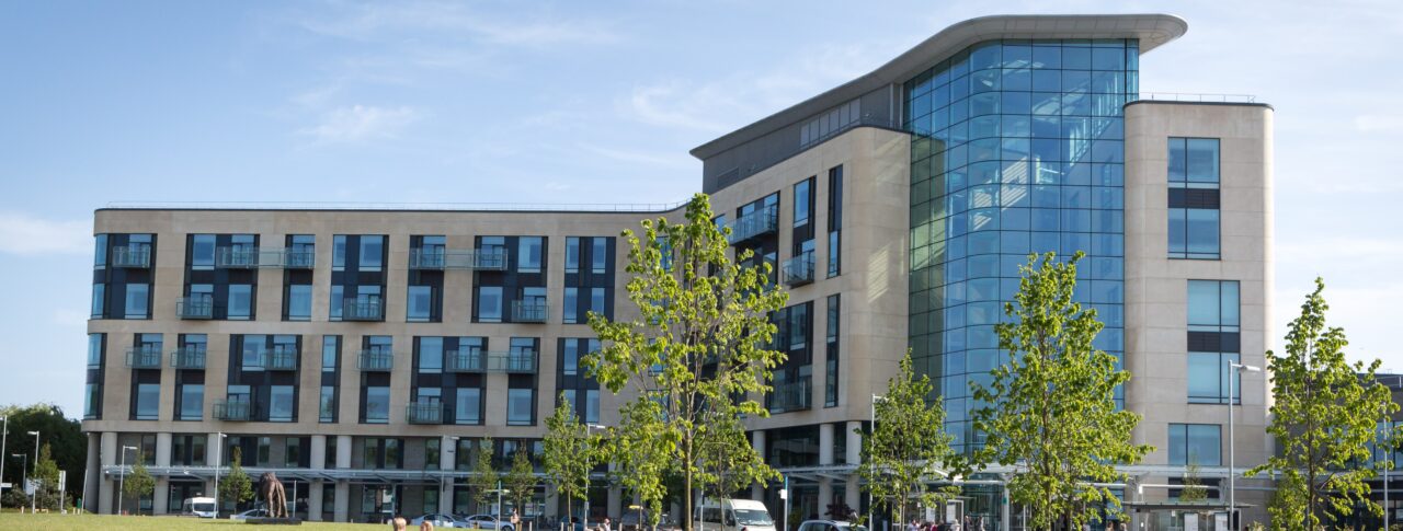 Southmead Hospital’s modern main building, featuring large glass panels and light‑coloured stone, stands behind a landscaped green area with young trees. People are sitting on benches in the foreground, and cars and pedestrians are visible near the hospital entrance on a sunny day