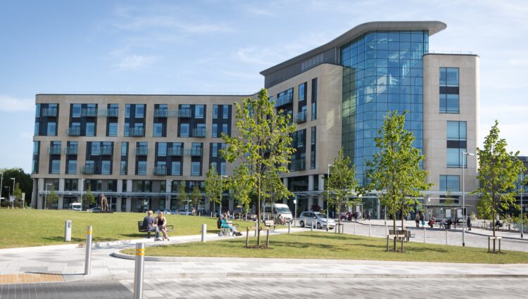 Southmead Hospital’s modern main building, featuring large glass panels and light‑coloured stone, stands behind a landscaped green area with young trees. People are sitting on benches in the foreground, and cars and pedestrians are visible near the hospital entrance on a sunny day